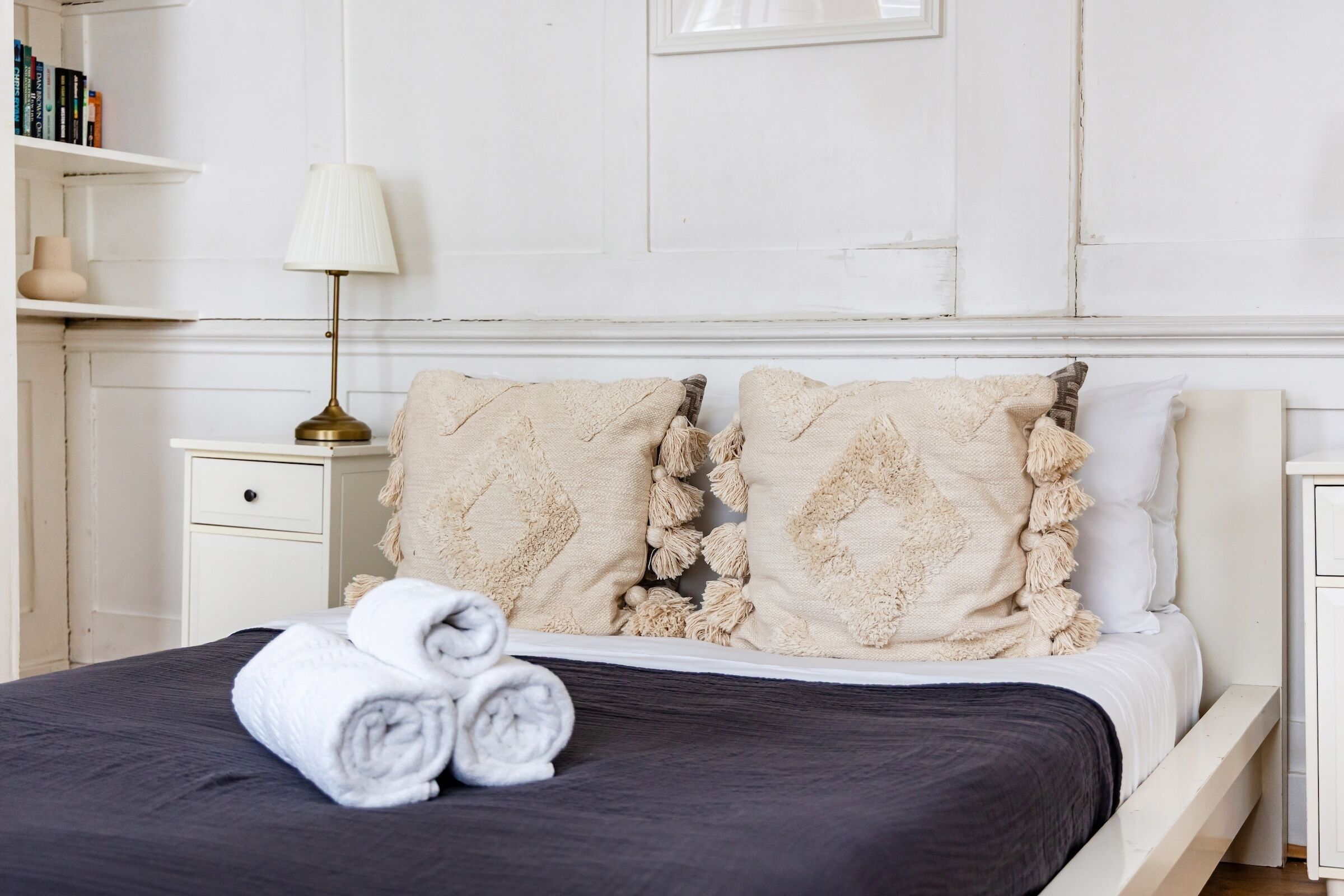 A neatly made bed with rolled towels, a bedside lamp, and a bookshelf with books.
