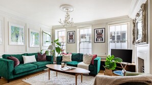 Ein Wohnzimmer mit einem grünen Sofa, einem Couchtisch und einem Fernseher. An den Wänden hängen Bilder in Rahmen und eine Deckenleuchte.
