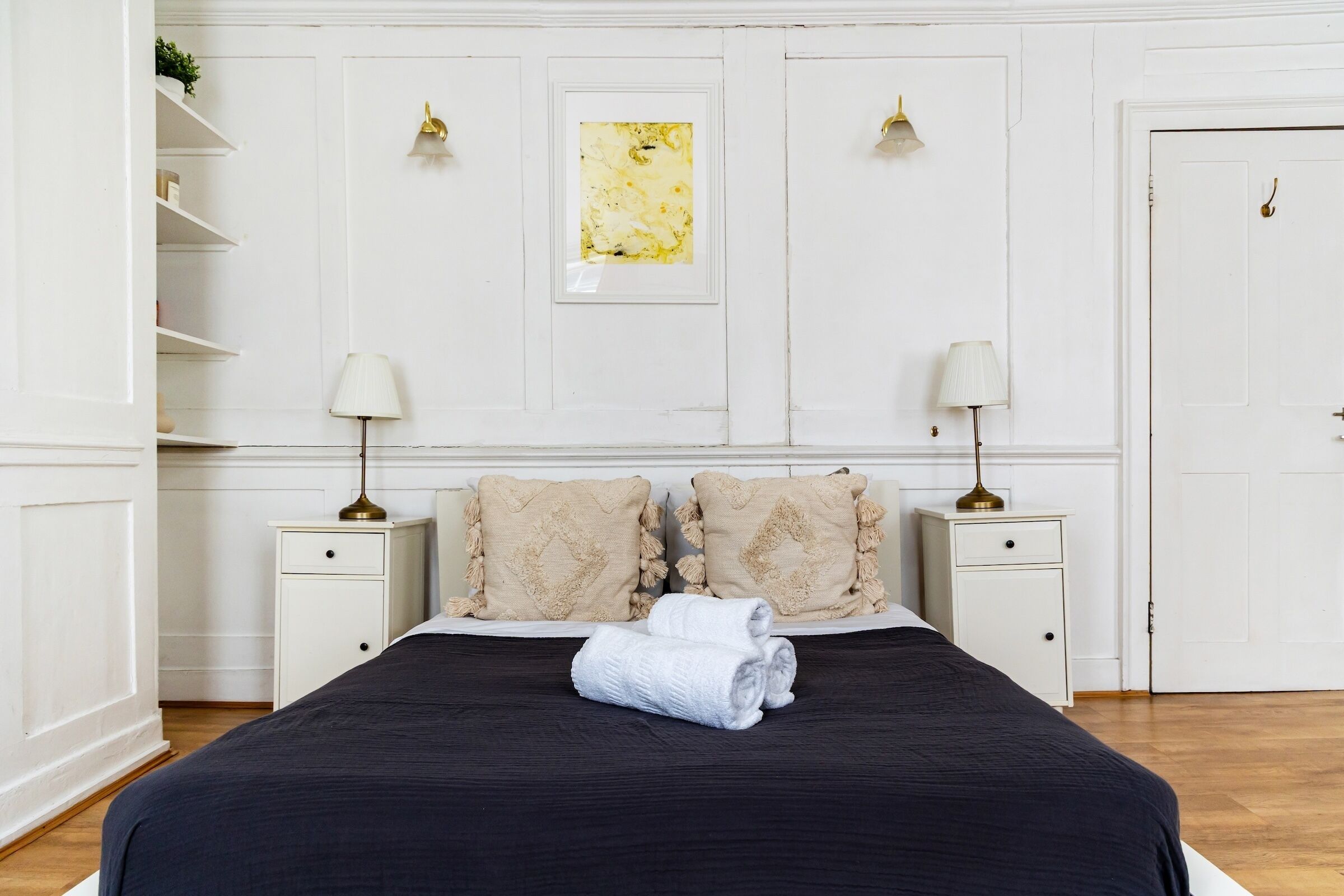 A neatly made bed with a dark bedspread and two pillows, flanked by white nightstands with lamps, in a room with white walls and a wooden floor.