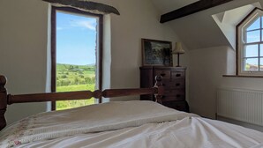 Un dormitorio con cama de madera, tocador, un cuadro y vista a un paisaje verde a través de un ventanal grande.
