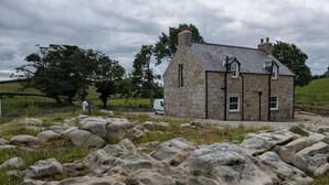 Una casa de piedra con techo inclinado, rodeada de grandes rocas y un pequeño estanque.