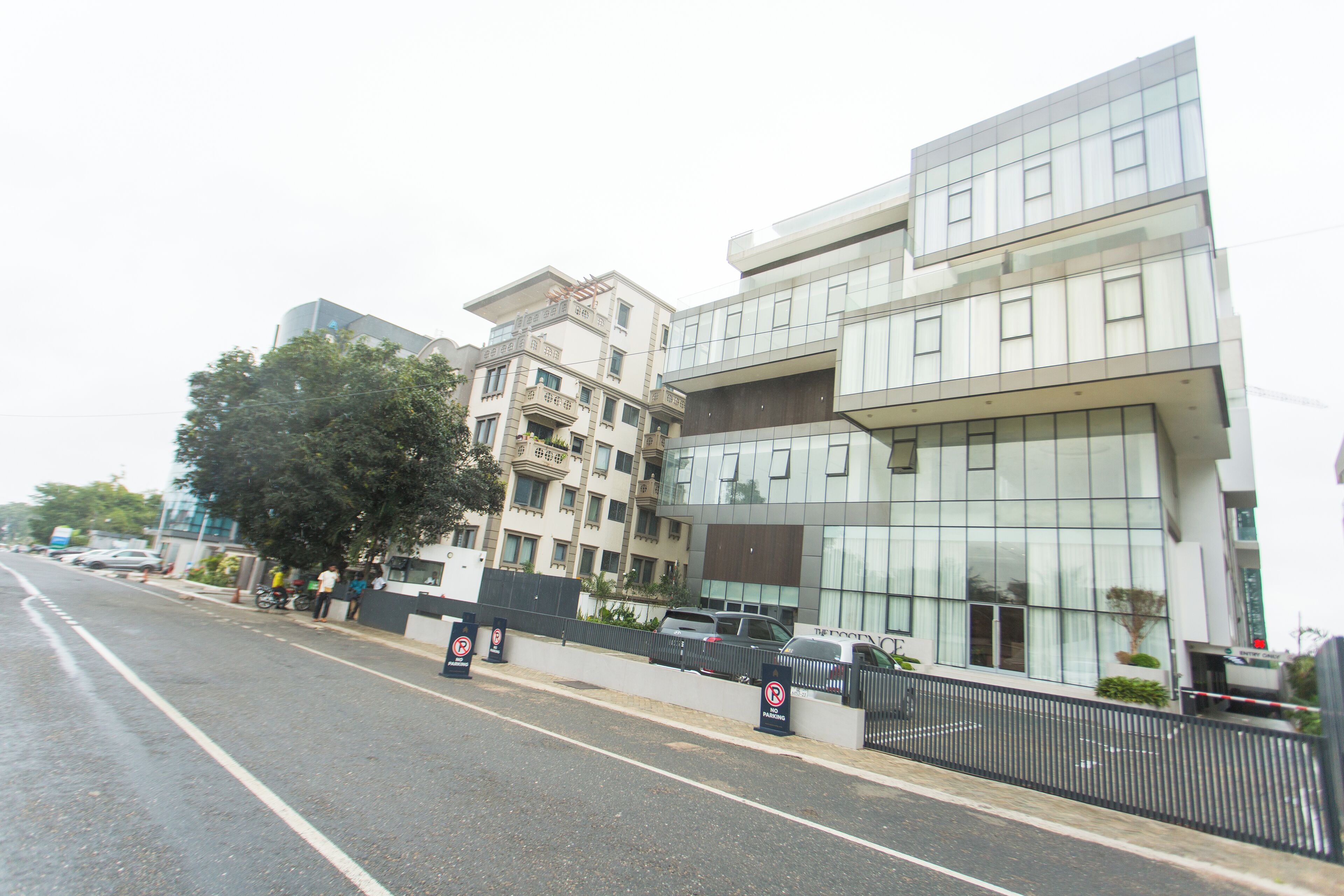 A modern building with a glass facade, a parking area, and a sidewalk with people.