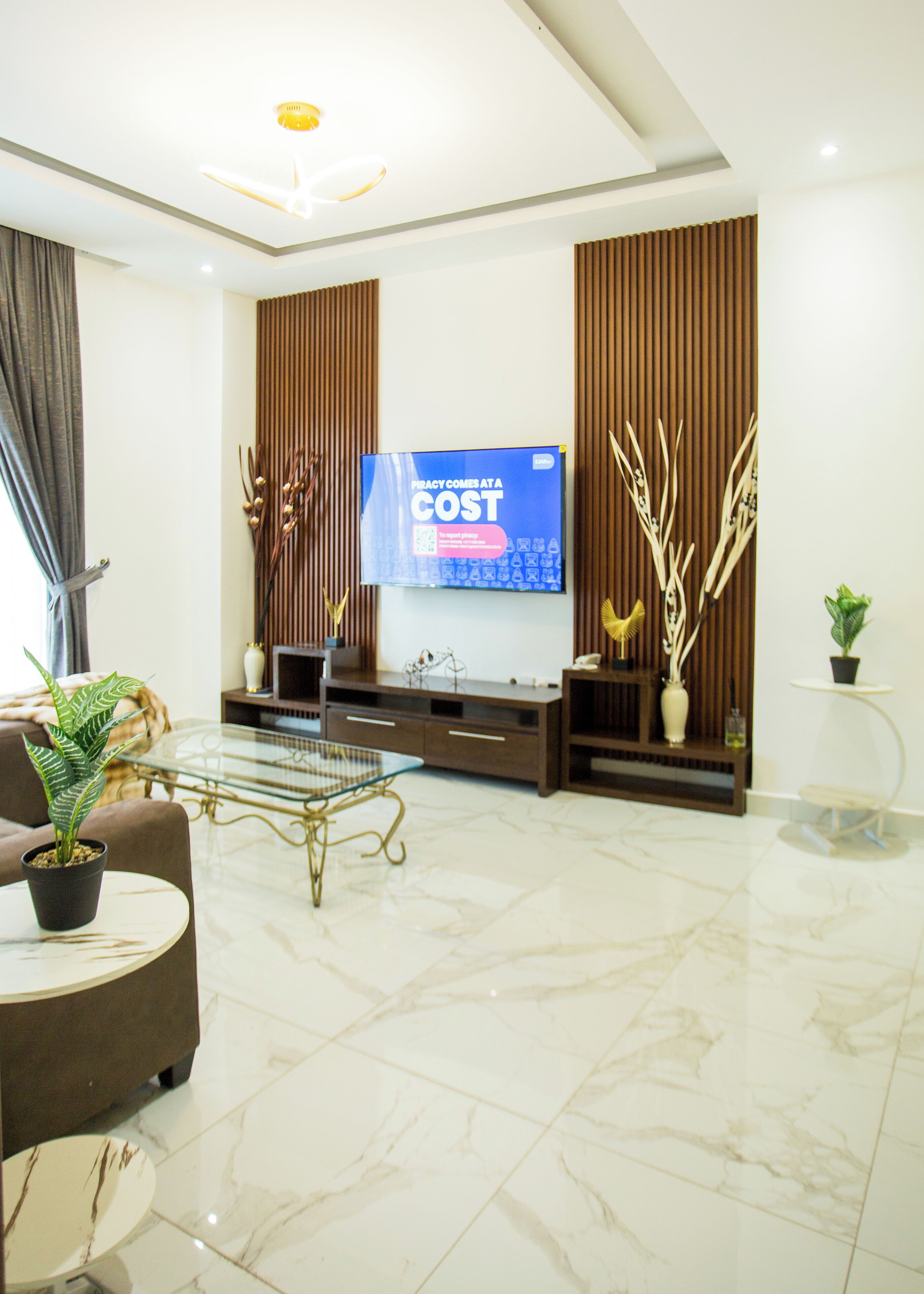 A modern living room with a large flat-screen TV mounted on the wall, a glass coffee table, a sofa, and decorative plants.