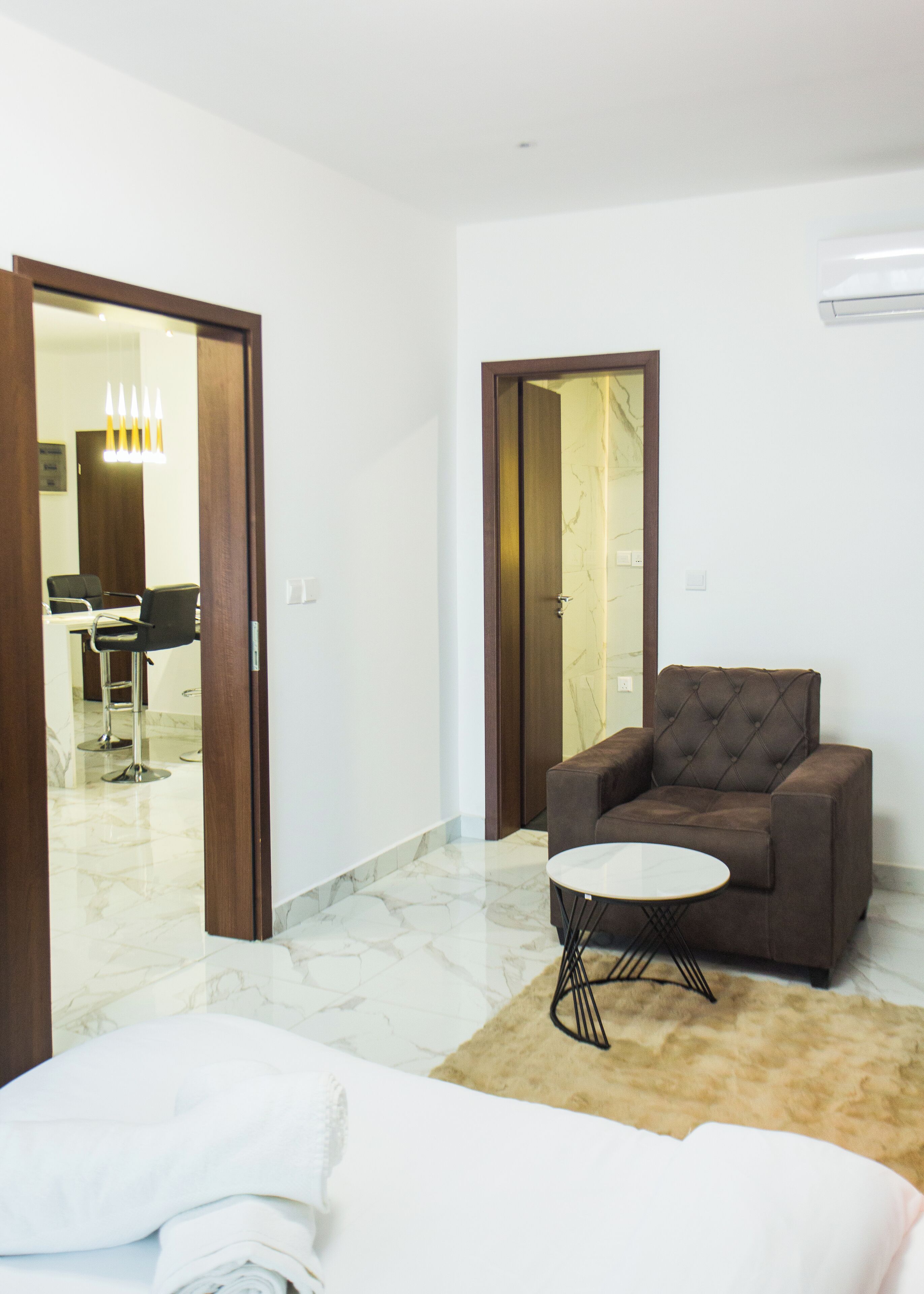 A modern hotel room with a marble floor, a brown sofa, a small round table, and a bed with a white blanket.