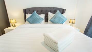 Una cama bien hecha con un cabecero tapizado gris, dos almohadas azules estampadas y una pila de toallas blancas. Hay dos lámparas de noche con pantallas a cada lado de la cama.