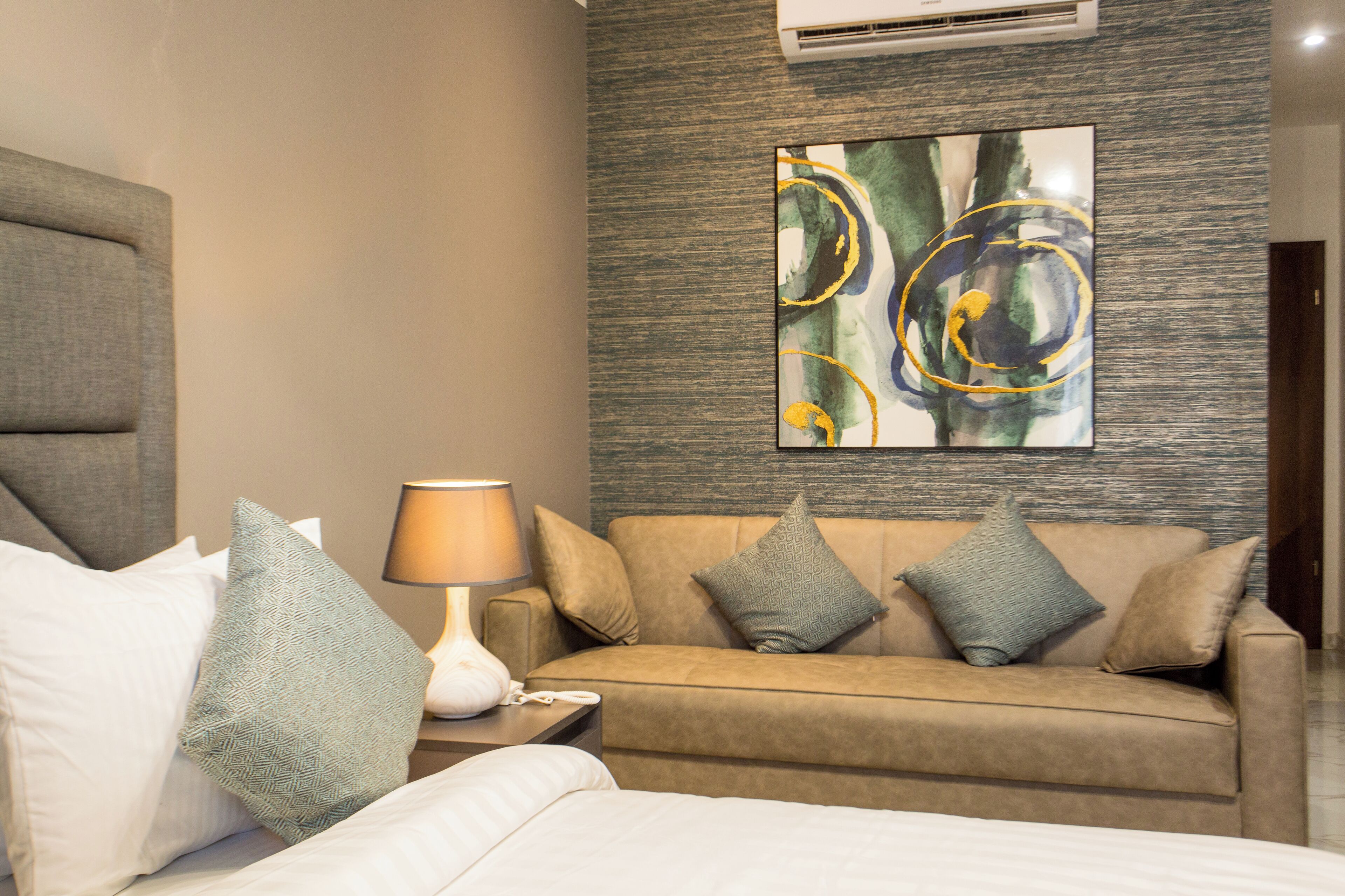 Habitación de hotel con cama, sofá, mesita de noche con lámpara y aire acondicionado instalado en la pared.