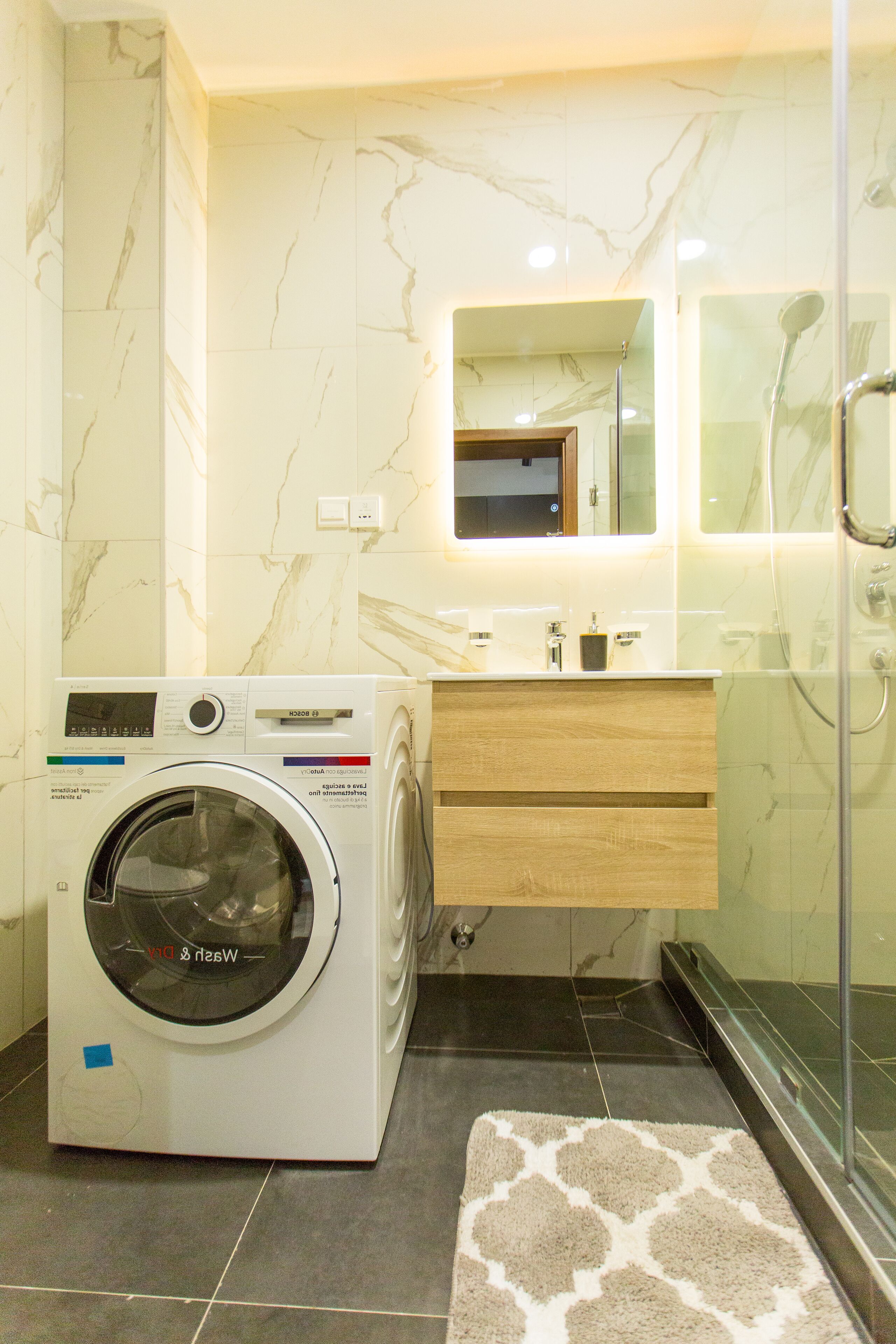 A modern bathroom with a built-in washing machine, a wooden vanity, a glass shower enclosure, and a marble-tiled wall.