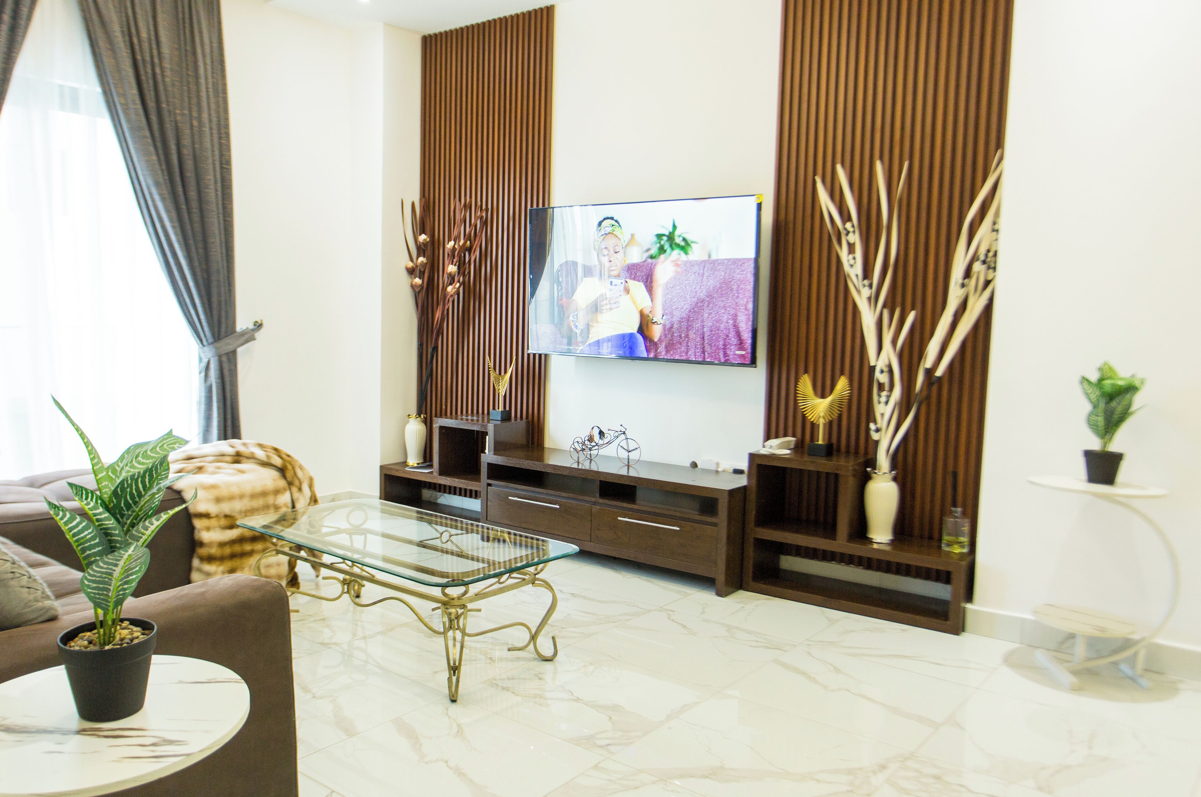 A modern living room with a glass coffee table, a sofa, a TV mounted on the wall, and decorative plants.