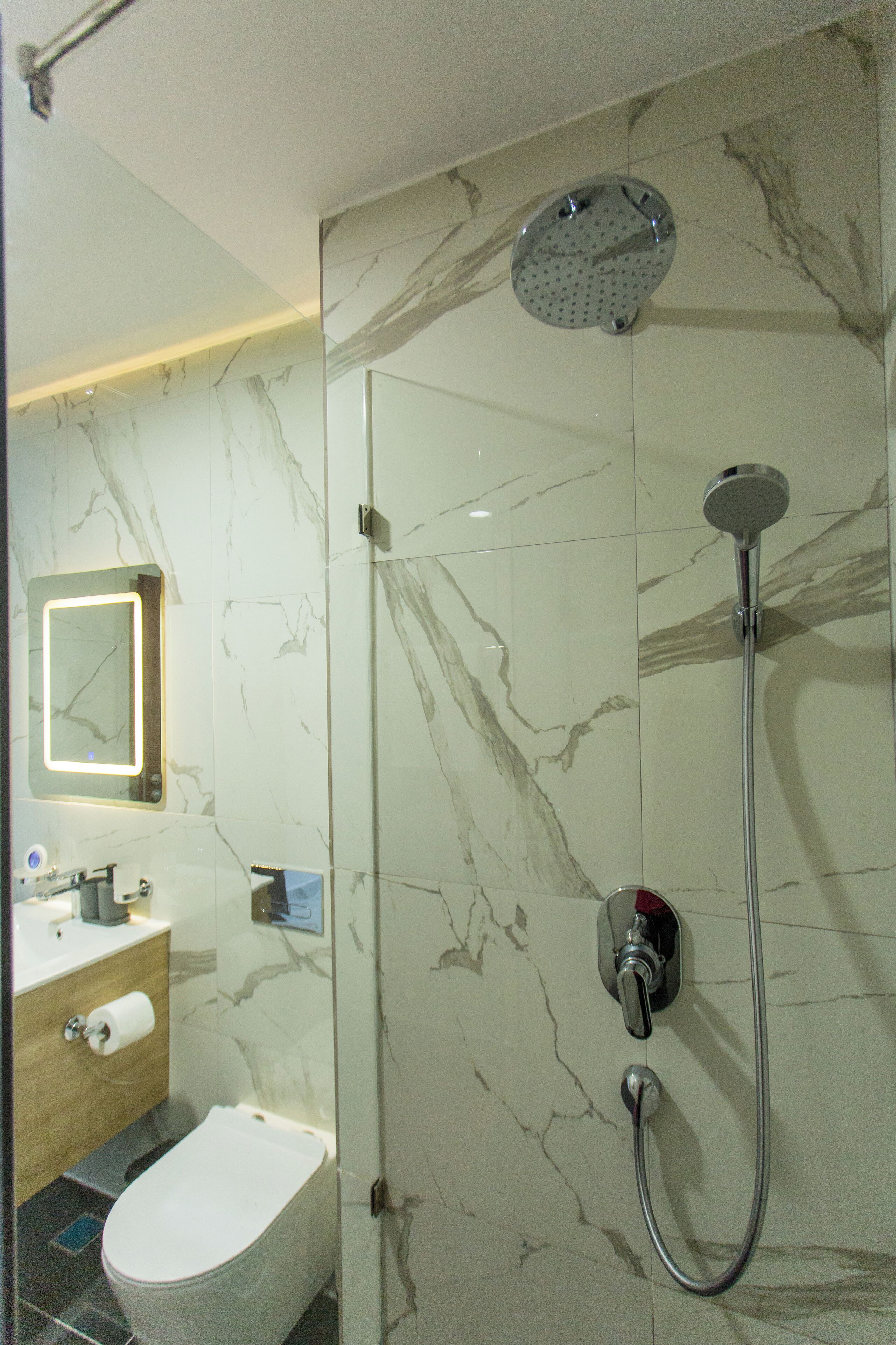 A modern bathroom with a marble wall, a toilet, a sink with a mirror, and a shower with a handheld showerhead.