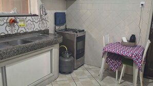 Cuina amb fogó de gas, rentapelats i frontissa de rajola. Hi ha un taulell amb mantell estampat i una cadira.