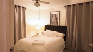 Une chambre d’hôtel avec un lit, une table de chevet, une lampe, un ventilateur de plafond et un tableau encadré au mur.