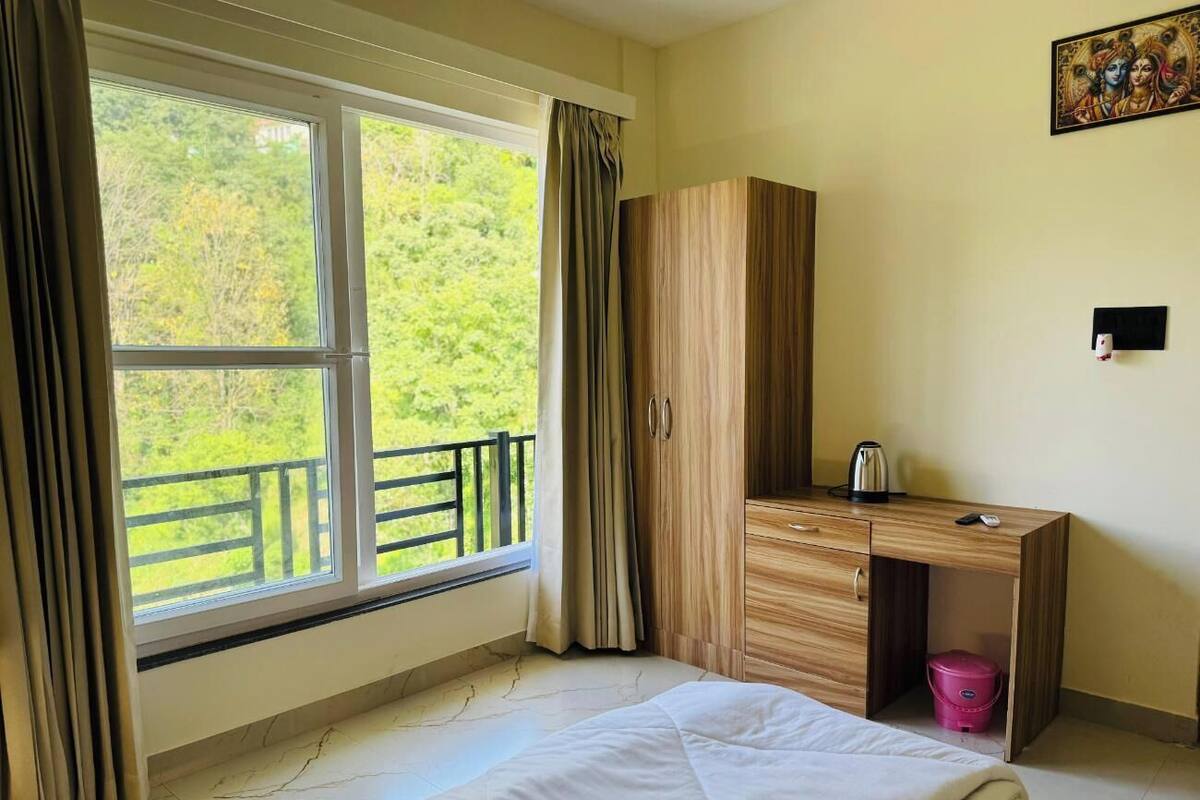 Quarto com cama, secretária de madeira, pequeno armário, janela com cortinas e vista para o verde lá fora.