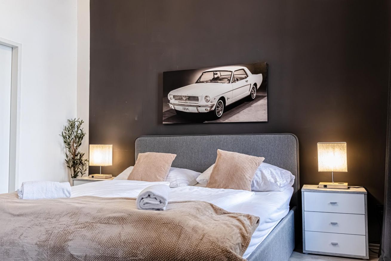Una habitación de hotel moderna con una cama grande, dos mesitas de noche con lámparas, un cuadro colgado en la pared de un coche clásico y una pequeña planta en maceta.