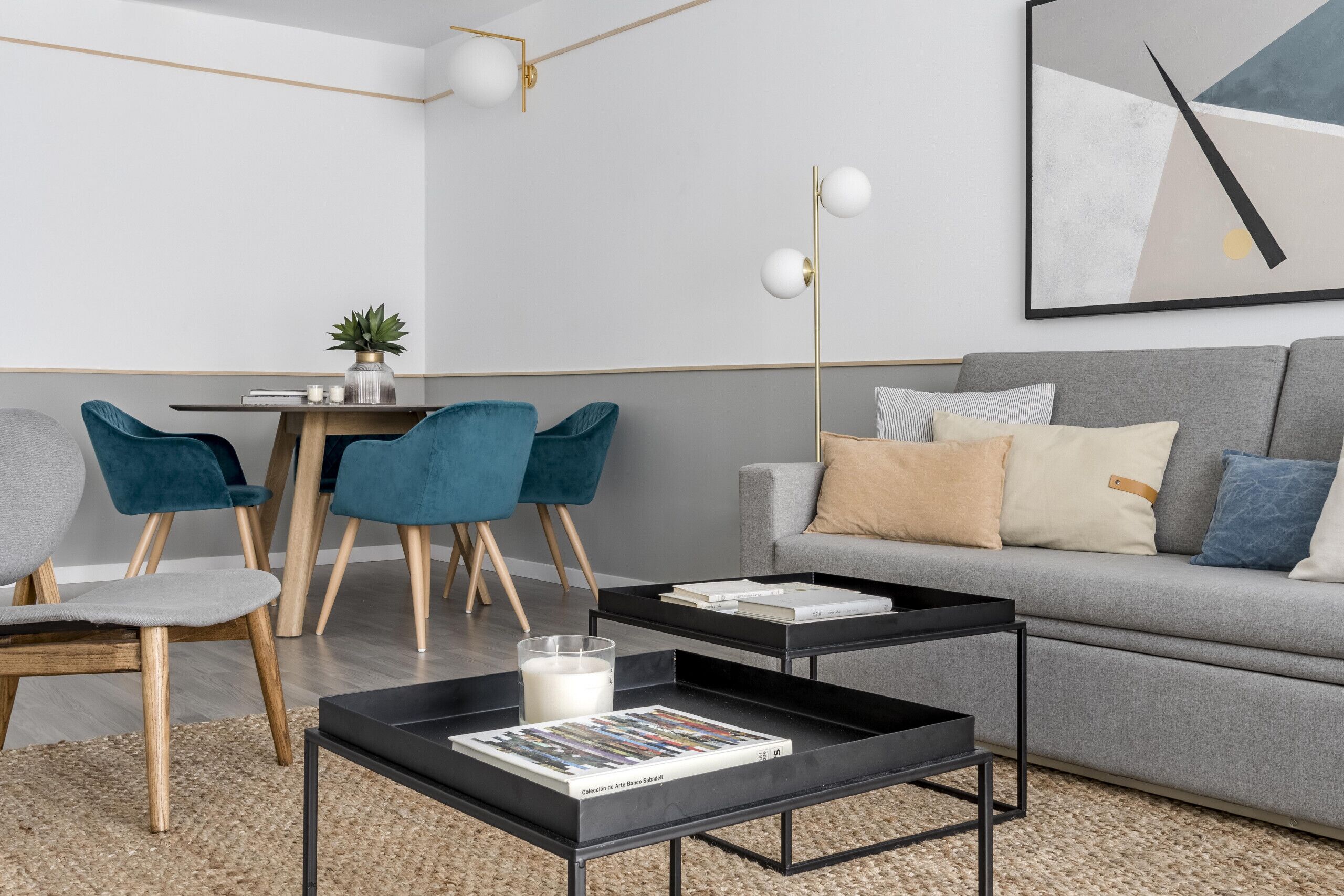 Una sala de estar moderna con un sofá gris, sillones turquesa, una mesa de centro con libros, una lámpara de pie y un cuadro en la pared.