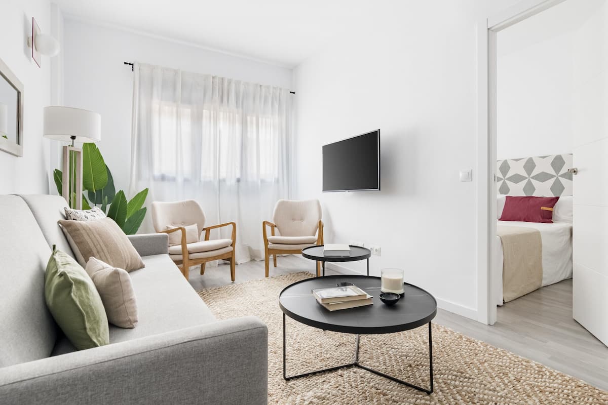 Una sala moderna con un sofá, dos sillones, una mesa de centro, un televisor y una cama con cabecera de estampado geométrico.