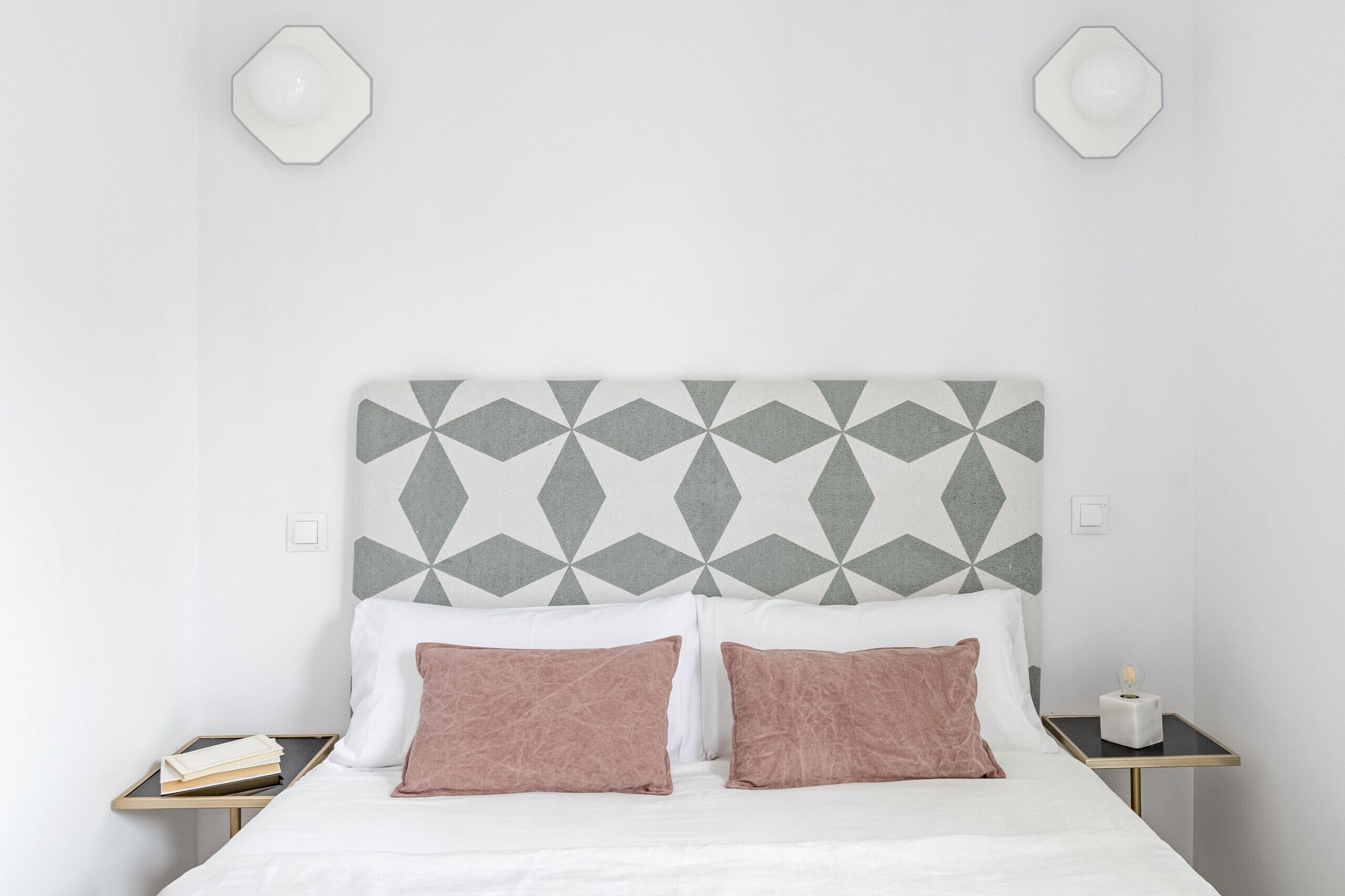 Un lit bien fait, avec une tête de lit à motifs géométriques, deux tables de chevet sur lesquelles reposent un livre et une boîte de mouchoirs, et un mur blanc uni.