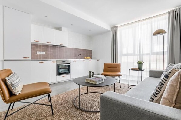 Una sala de estar moderna con un sofá gris, un sillón de cuero marrón y una mesa de centro. Se observa una zona de cocina con alacenas blancas y un revoque de azulejos.
