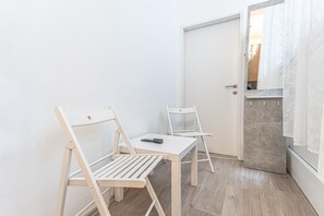 Ein kleiner, weißer Raum mit zwei Holzsesseln und einem Tisch, einer Tür und einem Spiegel, der einen Raum mit Vorhängen reflektiert.