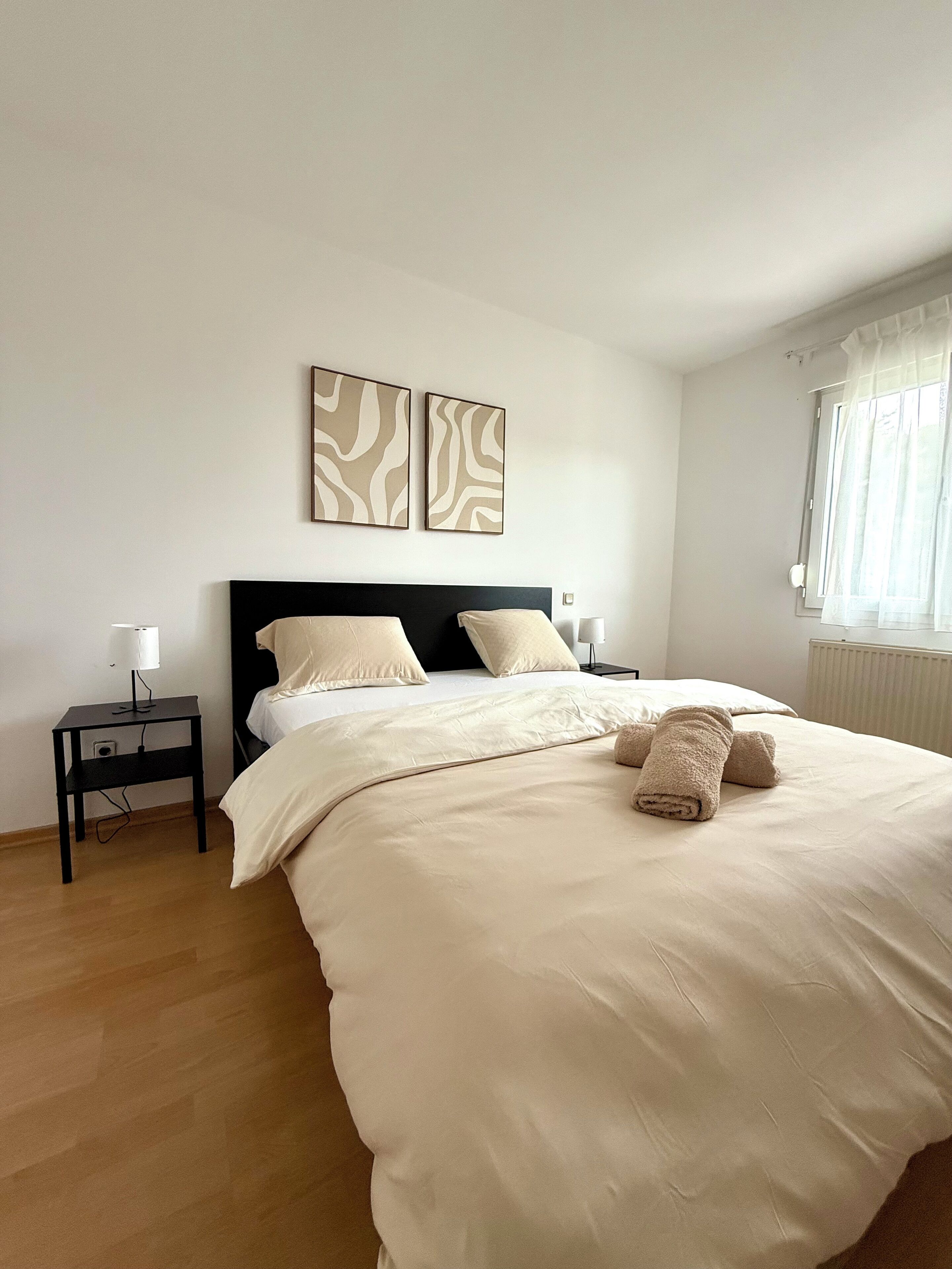 A neatly made bed with a beige comforter and pillows, a black nightstand with a lamp, and two wall-mounted abstract art pieces.