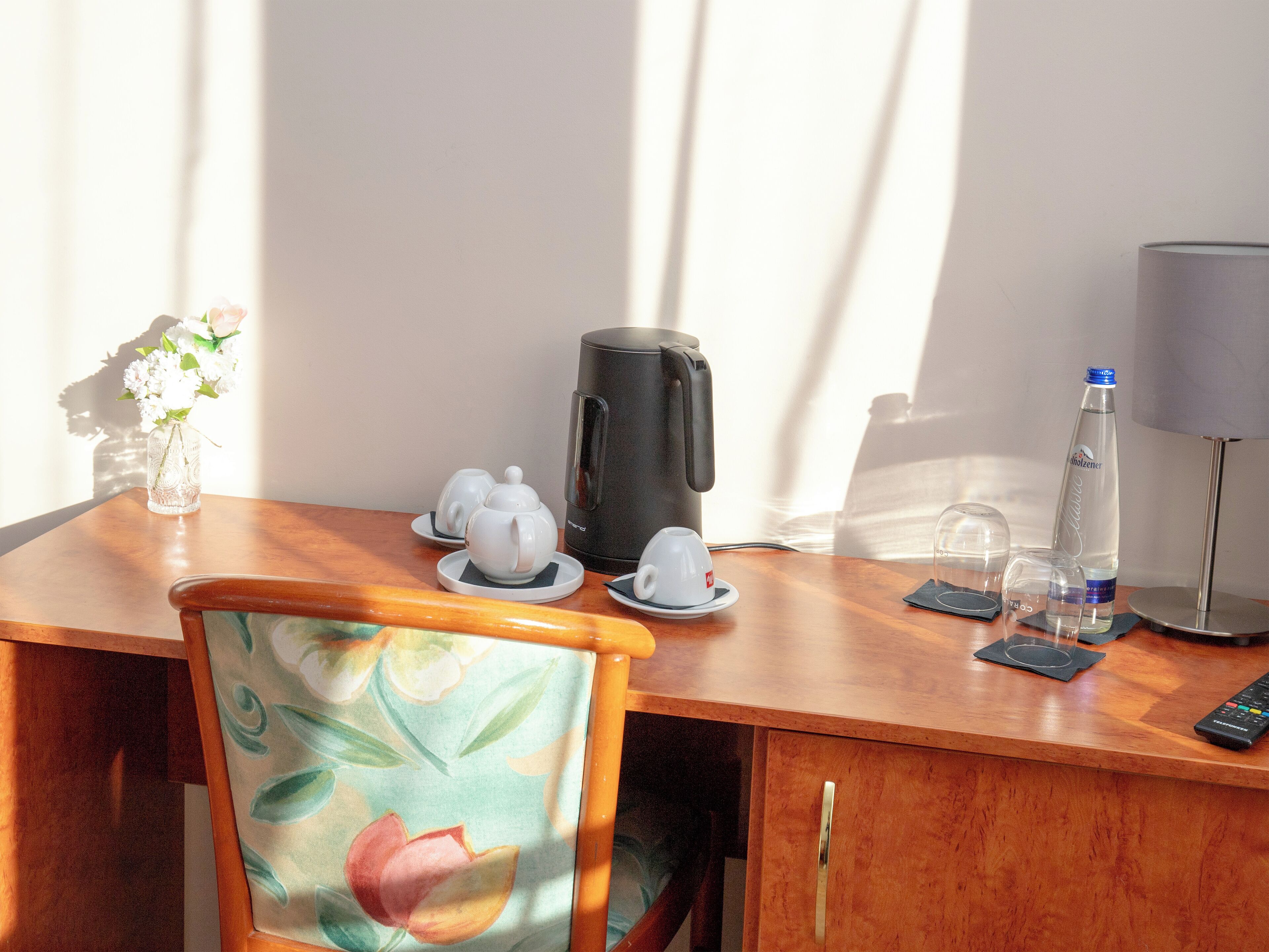 A wooden desk with a floral chair, a coffee maker, a water bottle, a glass, and a remote control.