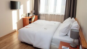 Una habitación de hotel con cama, mesita de noche con lámpara, una silla, televisor montado en la pared y ventana con cortinas.
