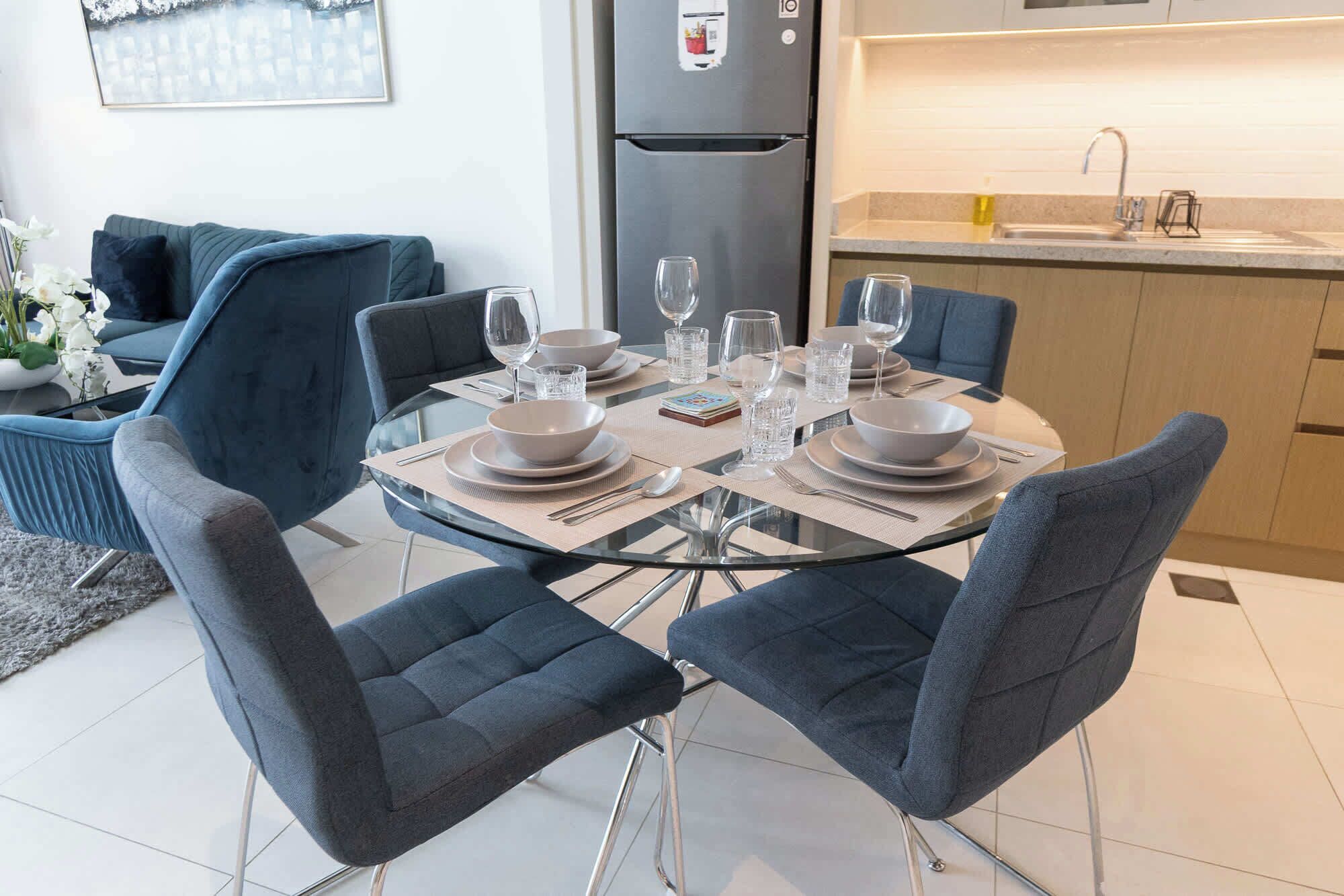 A modern dining area with a glass table set for four, surrounded by dark blue upholstered chairs. A kitchen with a stainless steel refrigerator and sink is visible in the background.