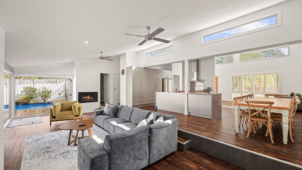 A spacious living area with a grey sectional sofa, a wooden dining table with chairs, a kitchen with white cabinets, and a pool visible through large windows.