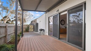 Un patio techado con cubierta de madera, una puerta corrediza de vidrio que da a una habitación con un reloj, y un jardín trasero cercado.