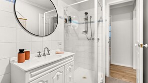Ein modernes Badezimmer mit einem weißen Waschtisch, einem runden Spiegel, einer Dusche inklusive Handbrause und einer kleinen Ablagefläche für Pflegeprodukte.