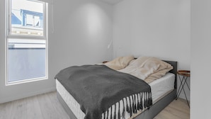 Un dormitorio con cama, mesita de noche, una pequeña mesa redonda y ventana con vistas a un edificio.