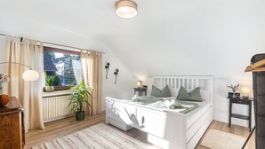 Un dormitorio con cama, mesita de noche, lámpara, radiador, ventana con cortinas y vistas a edificios.