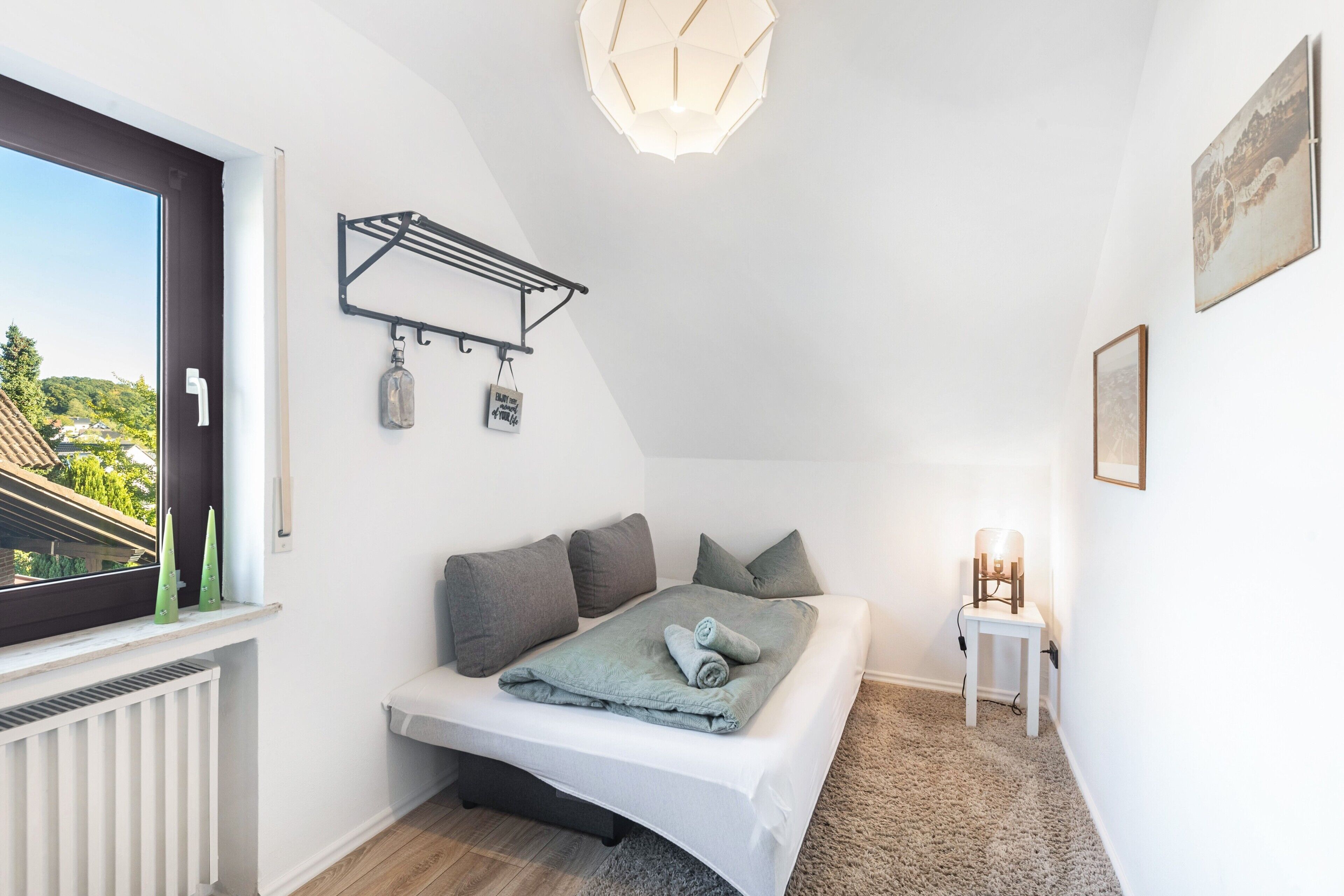A bedroom with a bed, a window, a radiator, a wall-mounted shelf, and a small table with a lamp.