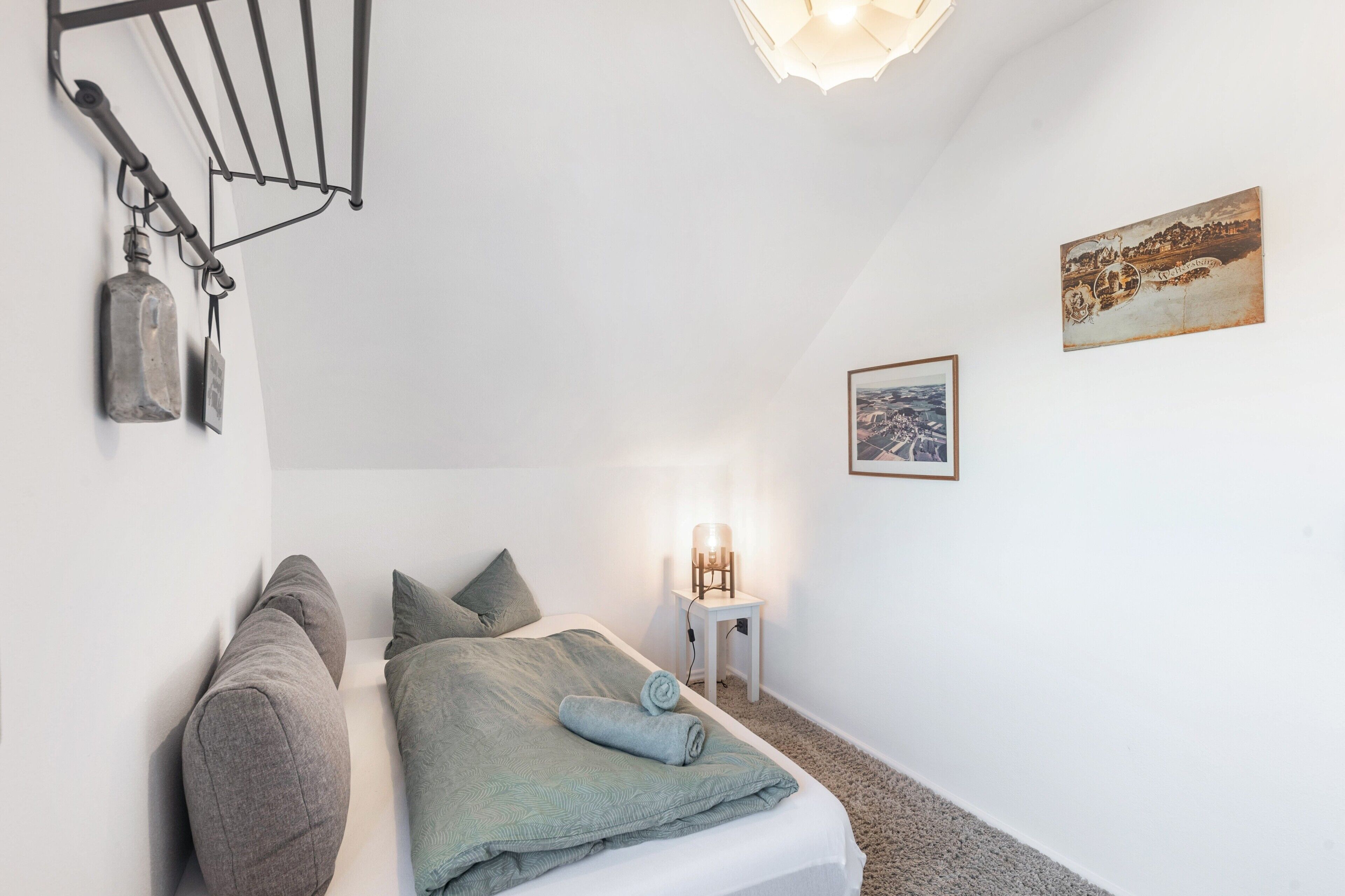 Un dormitorio pequeño y ordenado con cama, almohadas, una lámpara colgante, una mesita con lámpara y un cuadro en la pared.