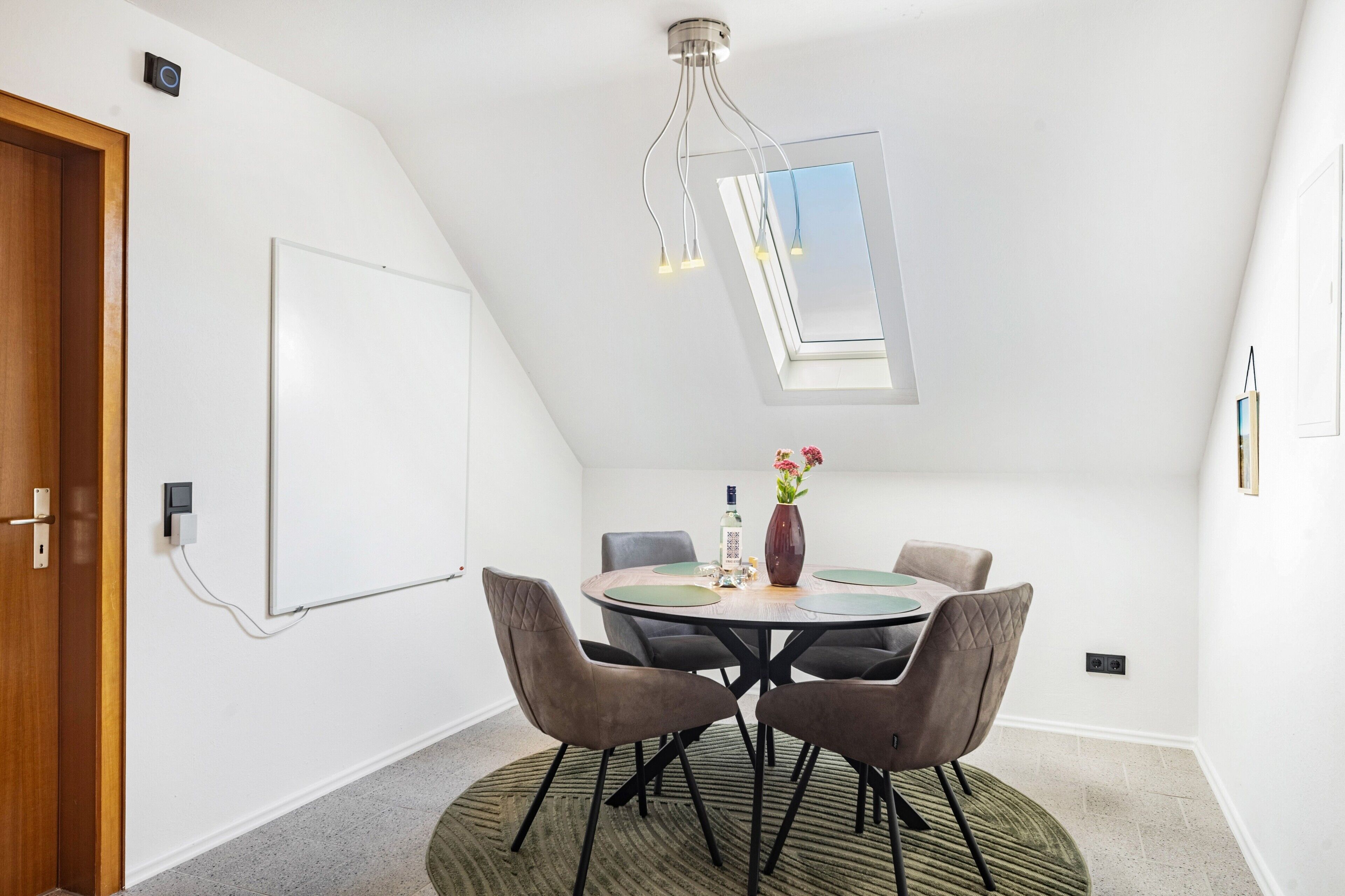A conference room with a round table, chairs, a vase with flowers, a whiteboard, and a skylight.