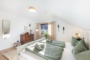 A bedroom with a sloped ceiling, a large window with curtains, a bedside table with a lamp, and a dresser.