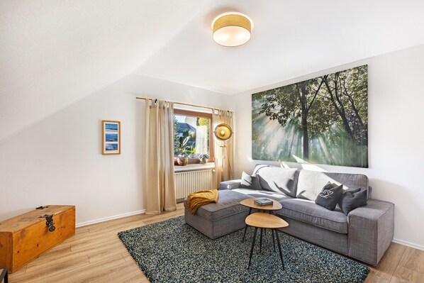 A modern living room with a grey sofa, a round coffee table, a large painting, a wooden chest, and a window with curtains.