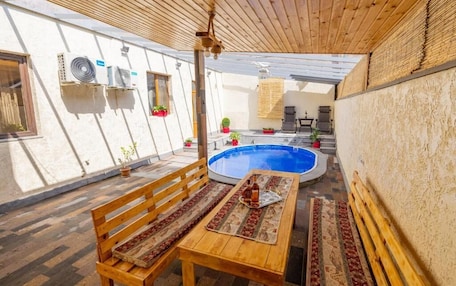 A covered outdoor area with a wooden bench, a small table with a bottle, and a pool.