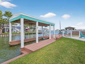 A covered outdoor area with a dock over water, a grassy area, and a fenced section with a chair.