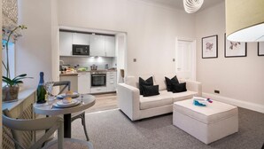 A modern living room with a white sofa, a dining table with chairs, a kitchen in the background, and a grey carpet. - Courtyard Apartments (Bowness-on-Windermere)