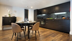 A modern kitchen with dark cabinetry, a tiled backsplash, and a dining area with two chairs and a table. - Courtyard Apartments (Bowness-on-Windermere)