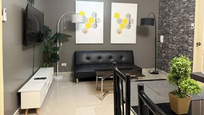 Ein modernes Wohnzimmer mit einem schwarzen Sofa, einem an der Wand montierten Fernseher, zwei gerahmten Kunstwerken, einer Topfpflanze und einer Treppe, die zu einer oberen Ebene führt.