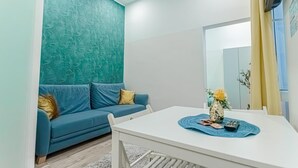 Una sala de estar moderna con una pared verde azulada estampada, un sofá azul, una mesa blanca con un florero y controles remotos, y una cortina amarilla.
