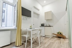 Una habitación pequeña y luminosa con una mesa y sillas blancas, un área de cocina con armarios y electrodomésticos, y una ventana con cortinas.