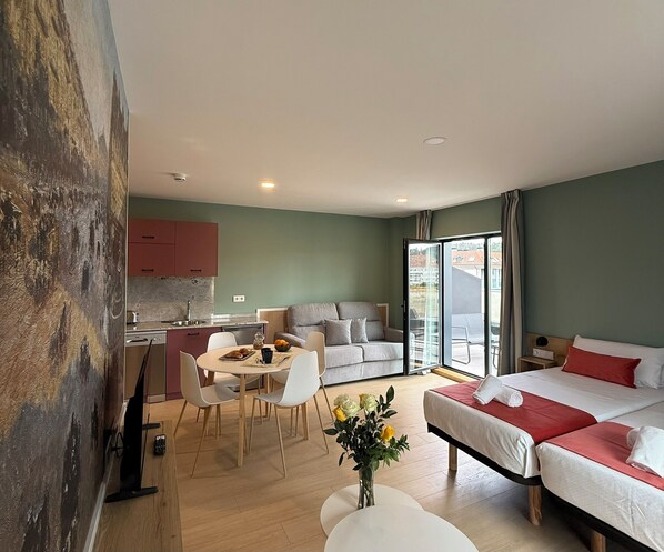 A modern hotel room with a dining area, kitchenette, and a large bed with red bedding.