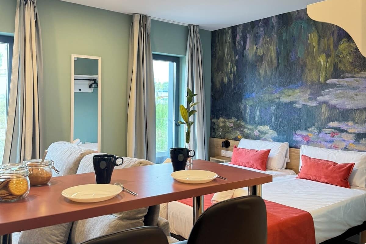A modern hotel room with a dining table, two beds, and a large abstract painting on the wall.