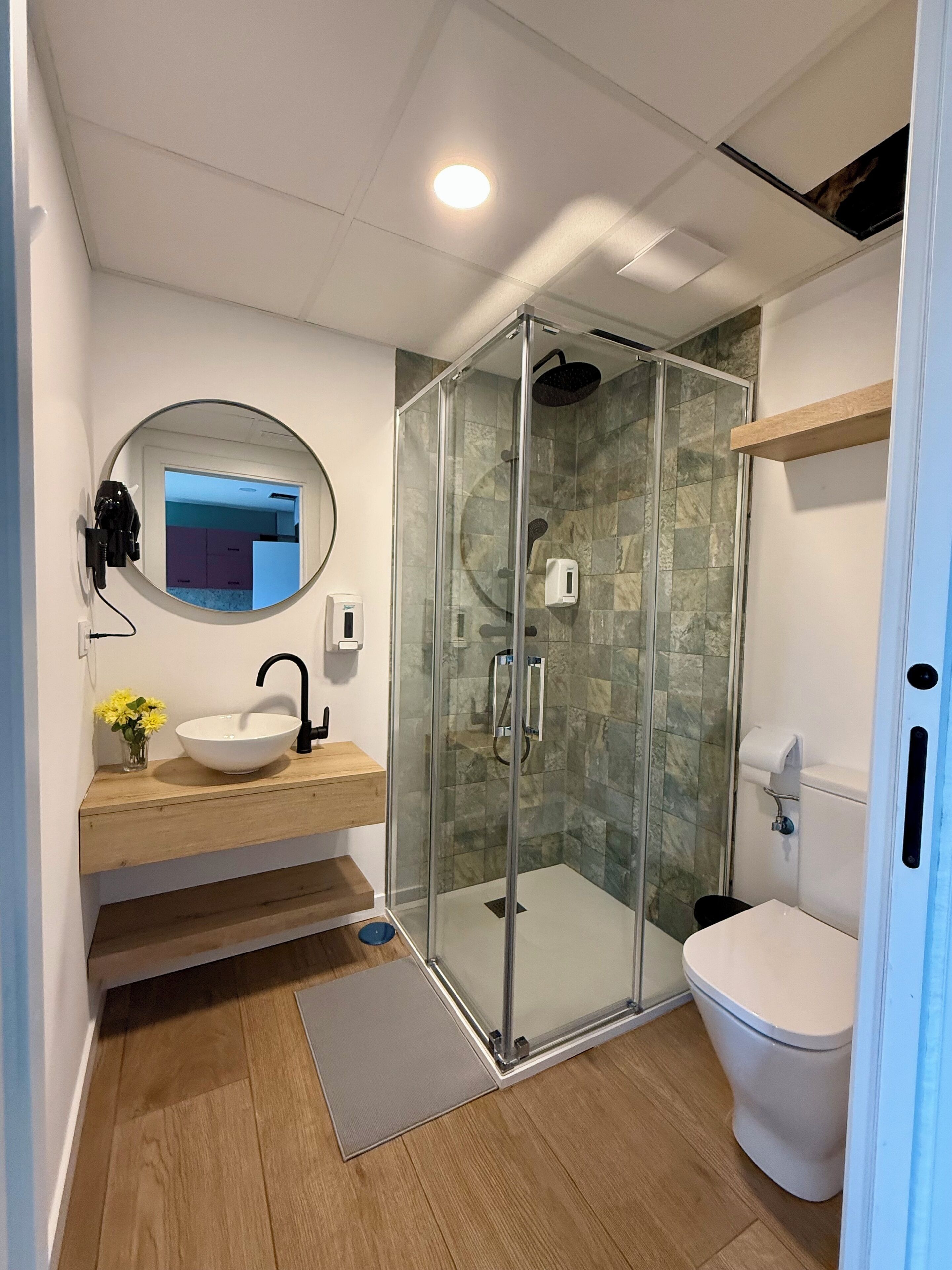 Ein modernes Badezimmer mit einer gläsernen Duschkabine, einem runden Spiegel, einem Holzkosmetikschrank und einer Toilette.