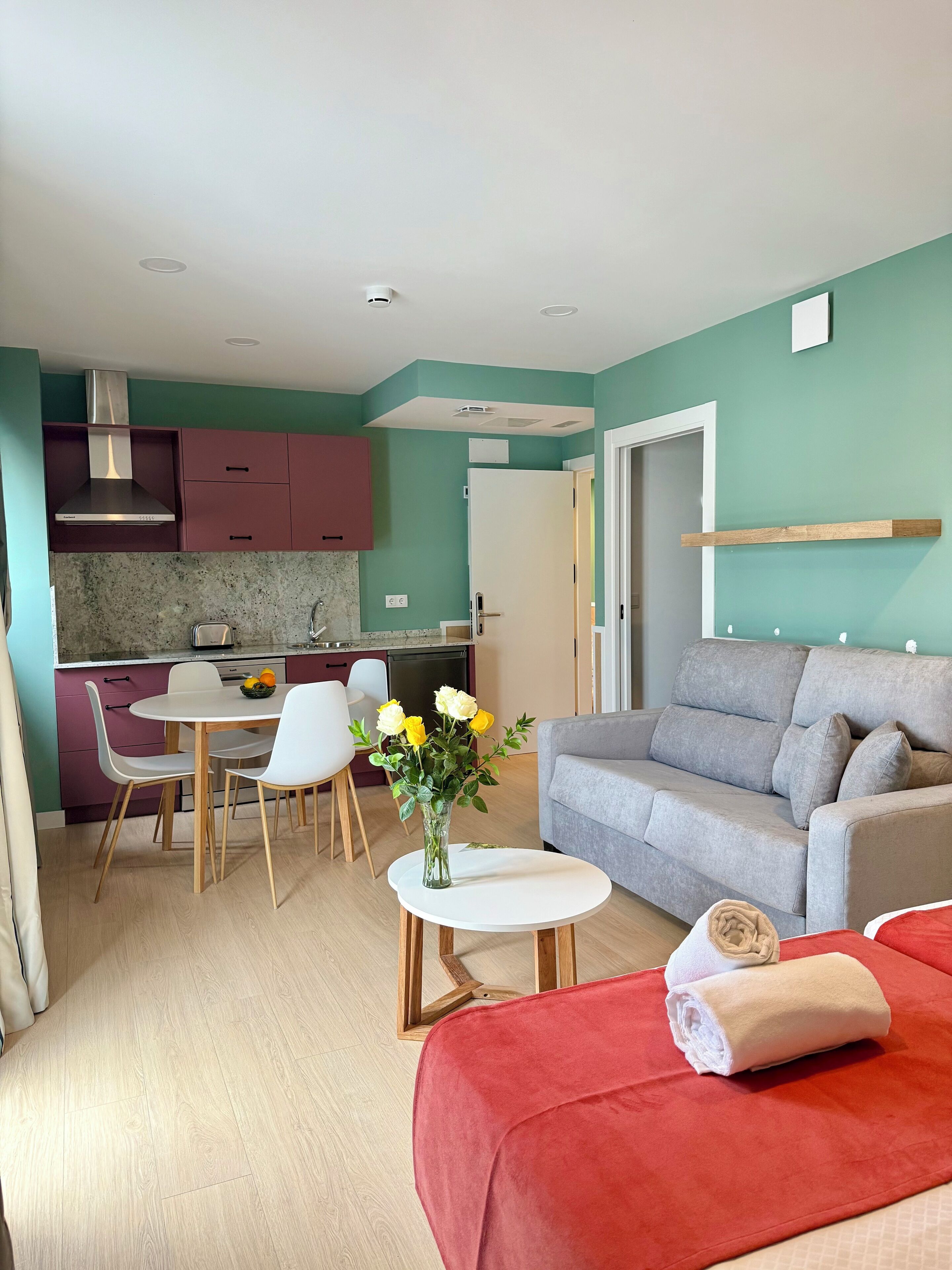A compact living space with a kitchenette, a dining area, and a seating area with a sofa and a red bed.