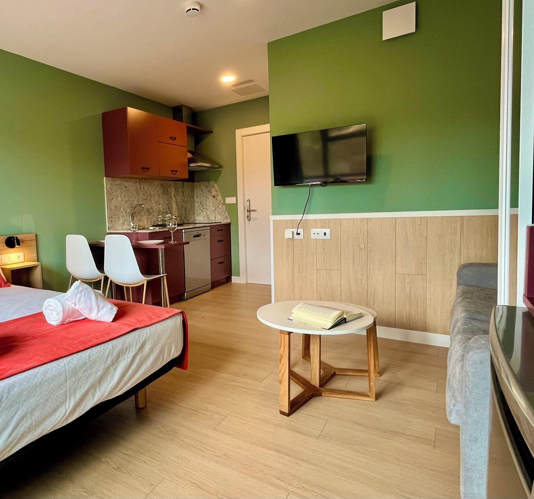A hotel room with a bed, a small table with a book, a TV mounted on the wall, a kitchenette, and a seating area with a couch.