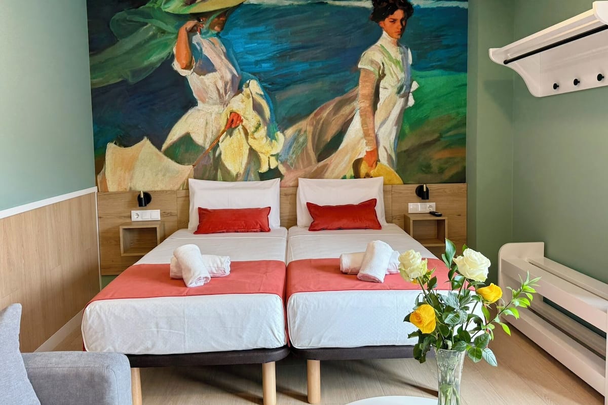 A hotel room with two beds, a large painting on the wall, a vase with flowers, and a white air conditioning unit.