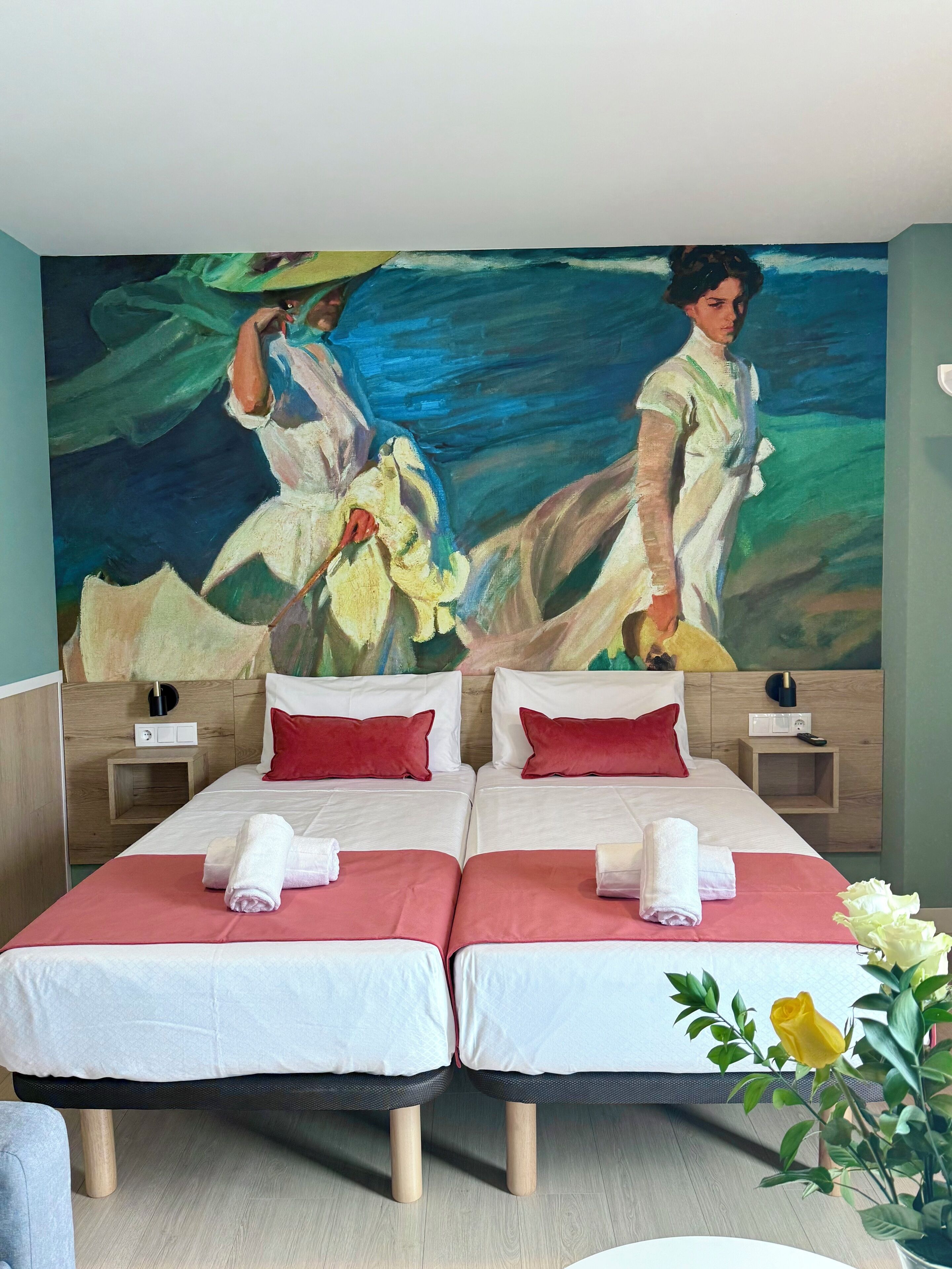 Habitación de hotel con dos camas, cada una con una almohada roja y una toalla blanca. En la pared hay un cuadro grande que representa a dos figuras con vestidos blancos.